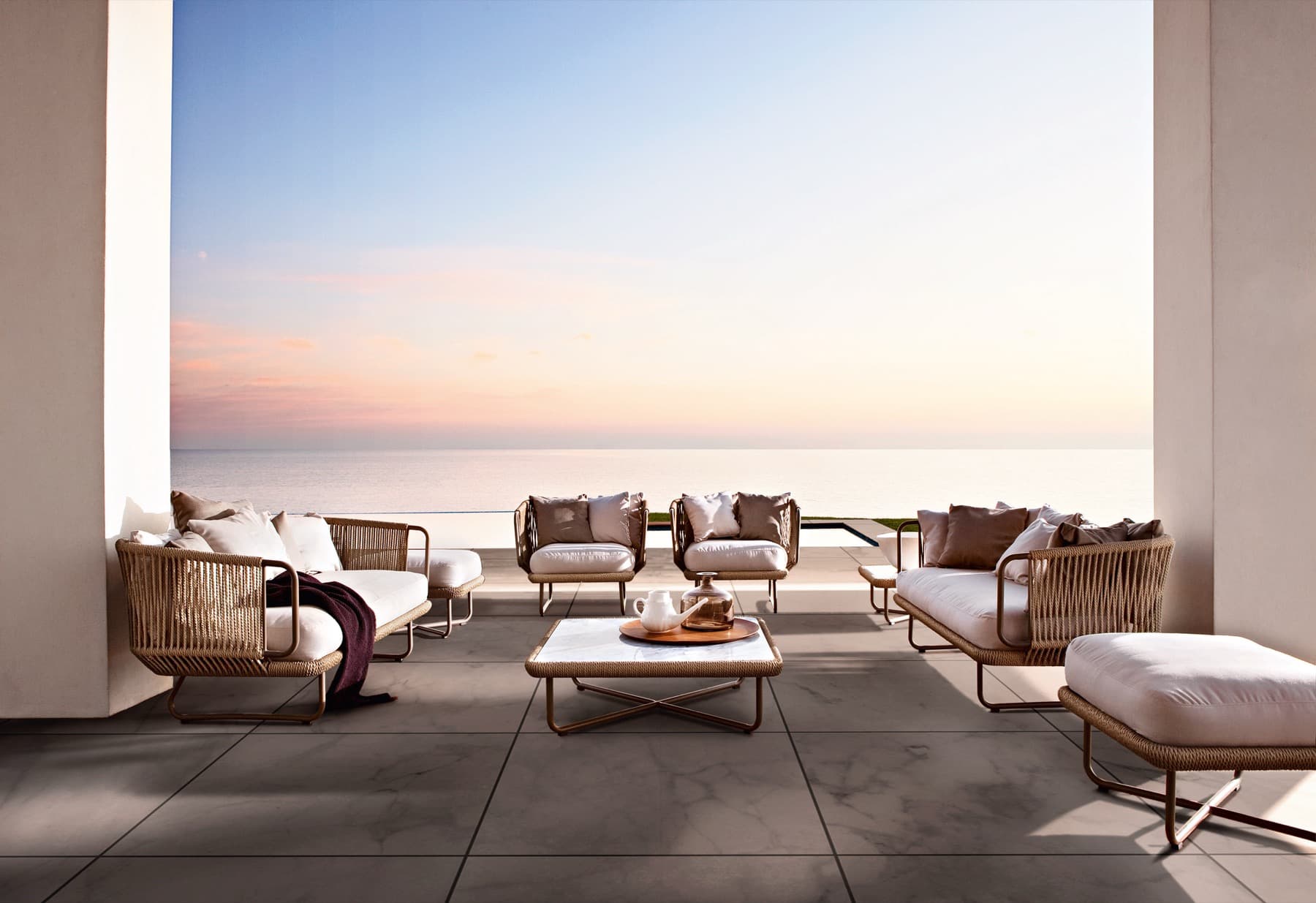 An outdoor terrace furnished with woven Varaschin lounge seating and a low table, looking out to the sea at sunset.